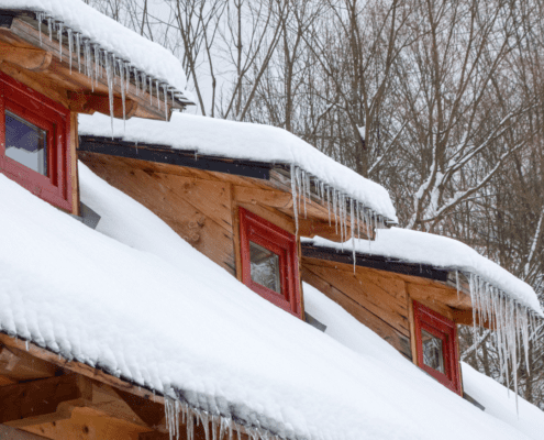 Snow and icicles on a wooden cabin roof with red windows.